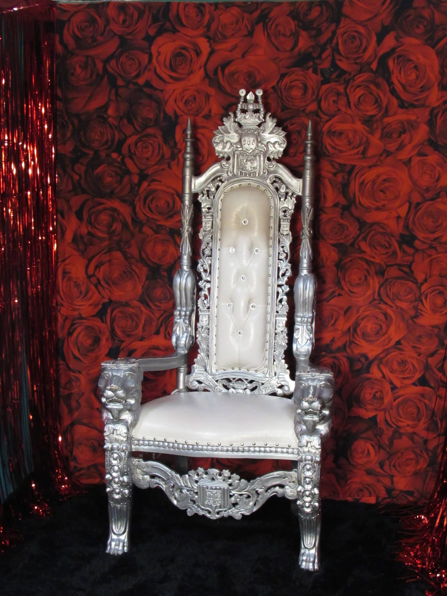 An ornate silver throne with detailed carvings and buttoned white upholstery set against a red rose patterned backdrop.
