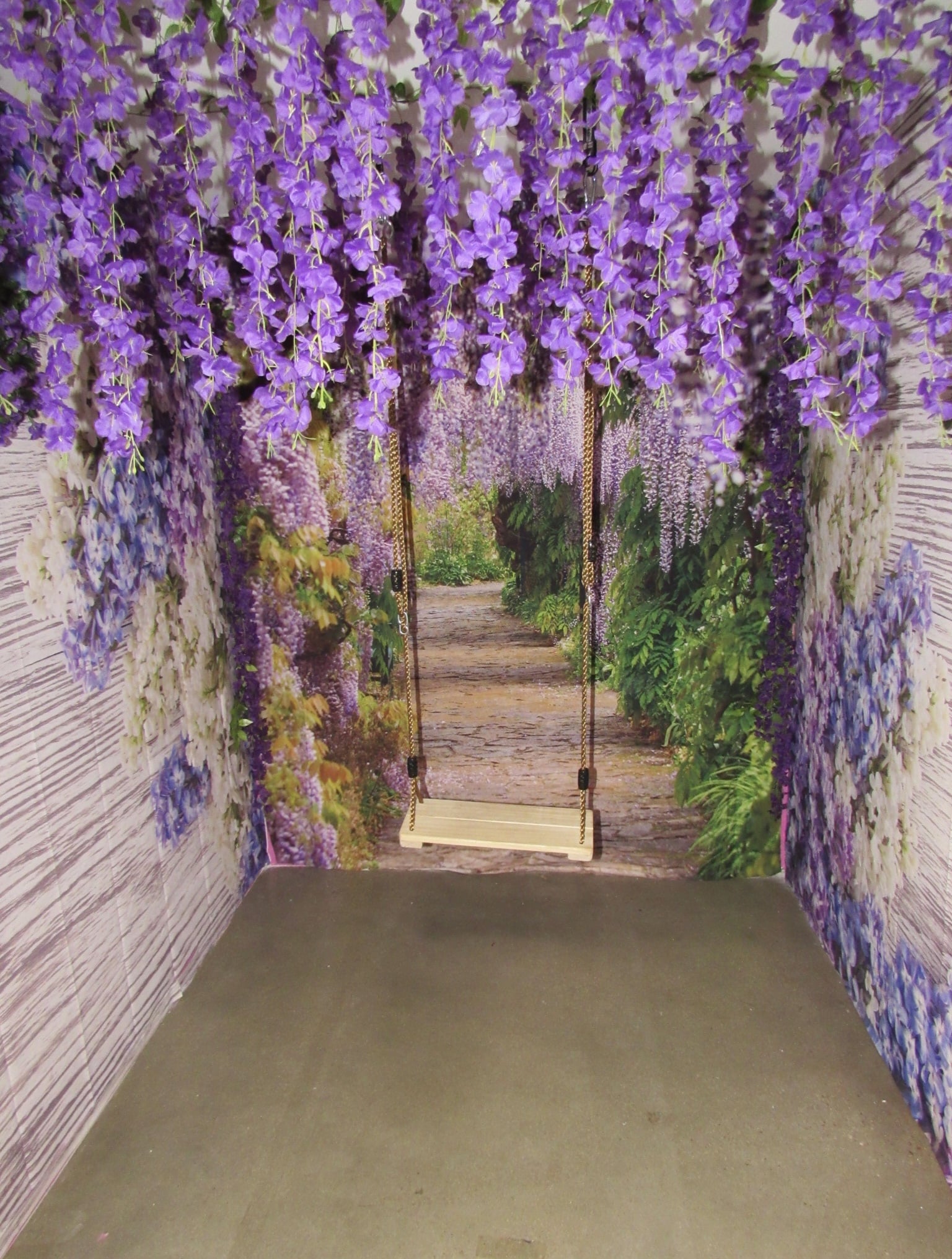 A whimsical indoor swing with purple wisteria flowers hanging from the ceiling and walls that show a forest path lined with lush greenery and flowers, creating a picturesque, fairy-tale atmosphere.
