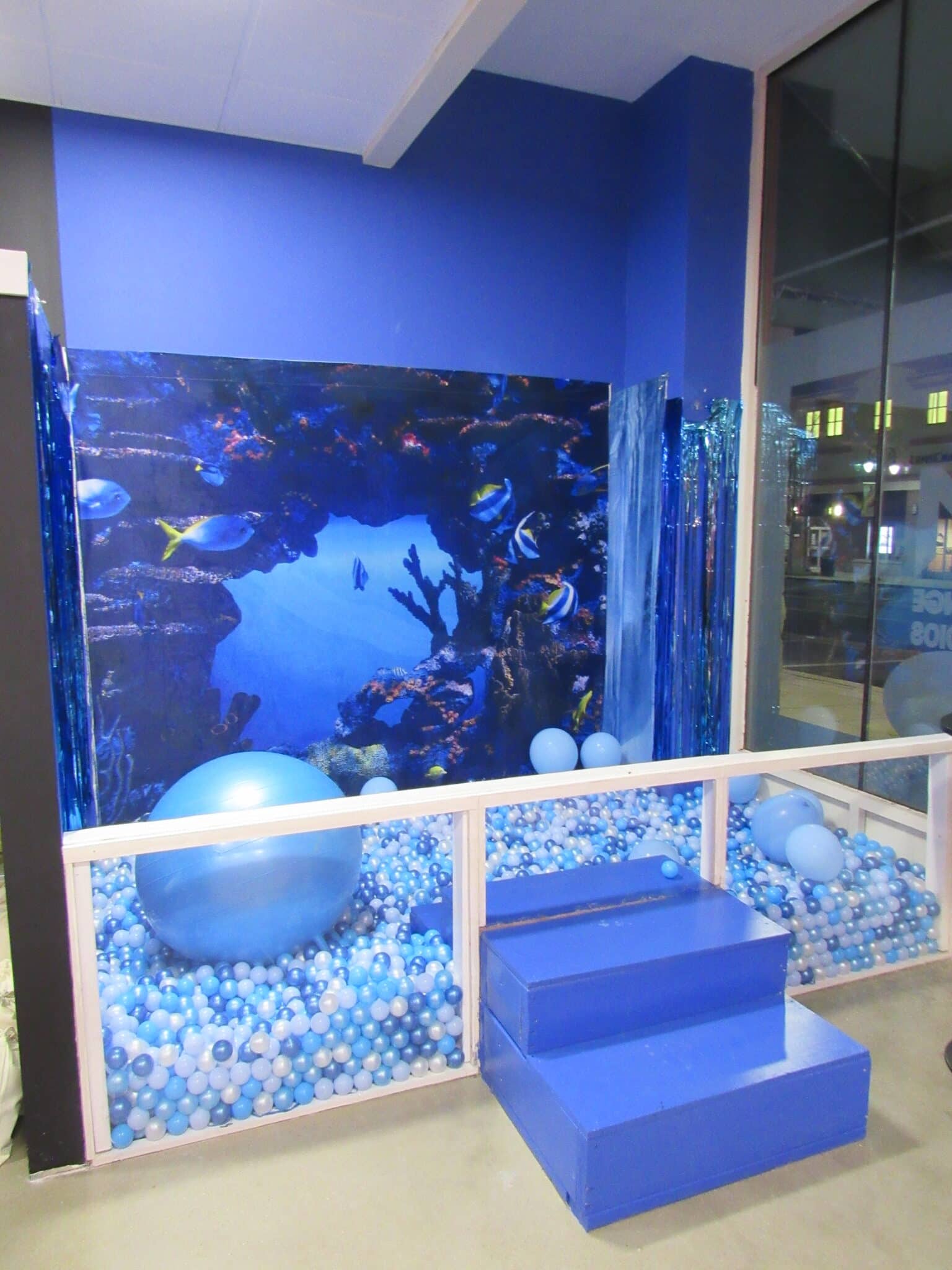 A vibrant office space featuring a large aquarium-themed mural with blue walls, assorted blue balloons, and steps leading up to the mural surrounded by a barrier.