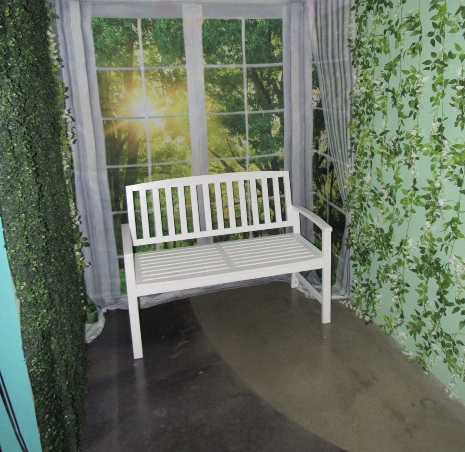 A white wooden bench sits in a staged indoor garden scene, surrounded by walls decorated with ivy patterns and a printed mural of a window overlooking a forest.
