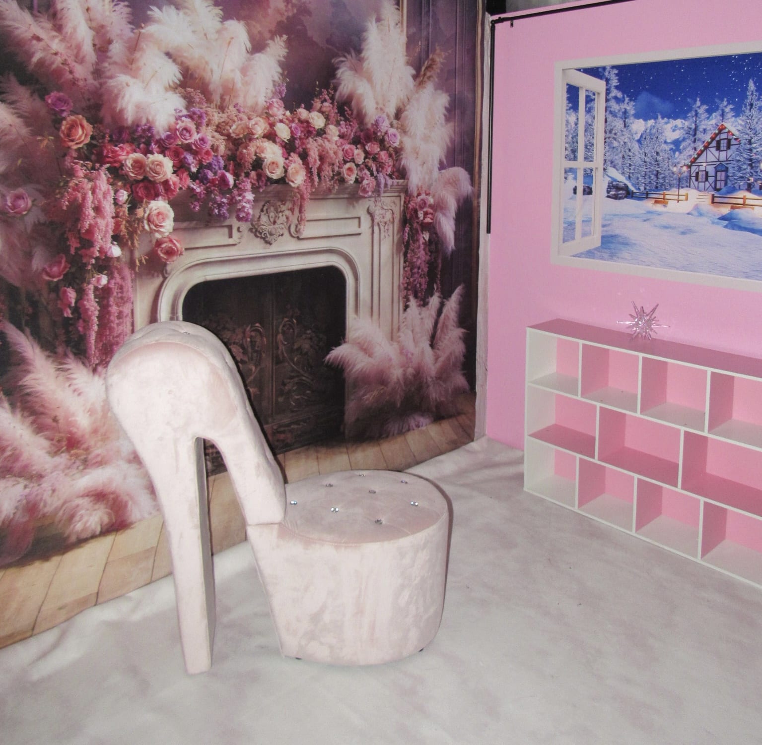 A whimsical indoor setting featuring an oversized plush pink shoe-shaped chair, a pink tiled counter, and walls adorned with floral and winter landscape images.