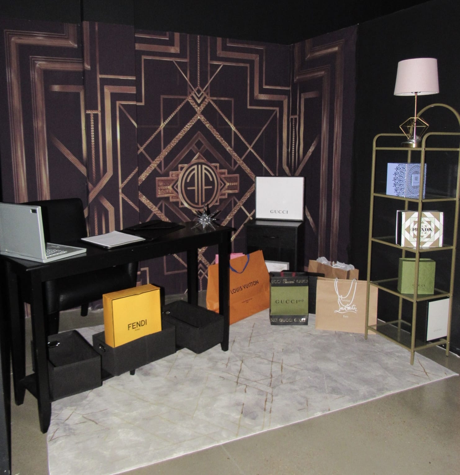 A chic luxury shopping display featuring a black desk with a laptop, surrounded by designer bags from fendi, gucci, and other brands, set against an art deco backdrop with a stylish lamp and shelf.