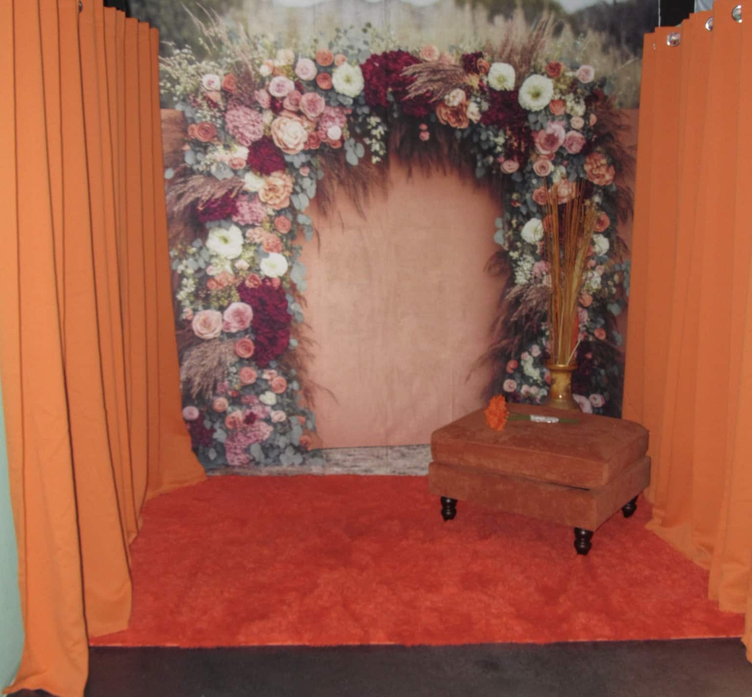 A cozy photo booth setup with an elaborate floral frame of mixed flowers, a small orange ottoman, and a vase of flowers on red carpeting, flanked by orange curtains.