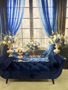 An elegant setup featuring a deep blue velvet sofa against a large window draped with translucent blue curtains, flanked by tall floral arrangements in gold vases.