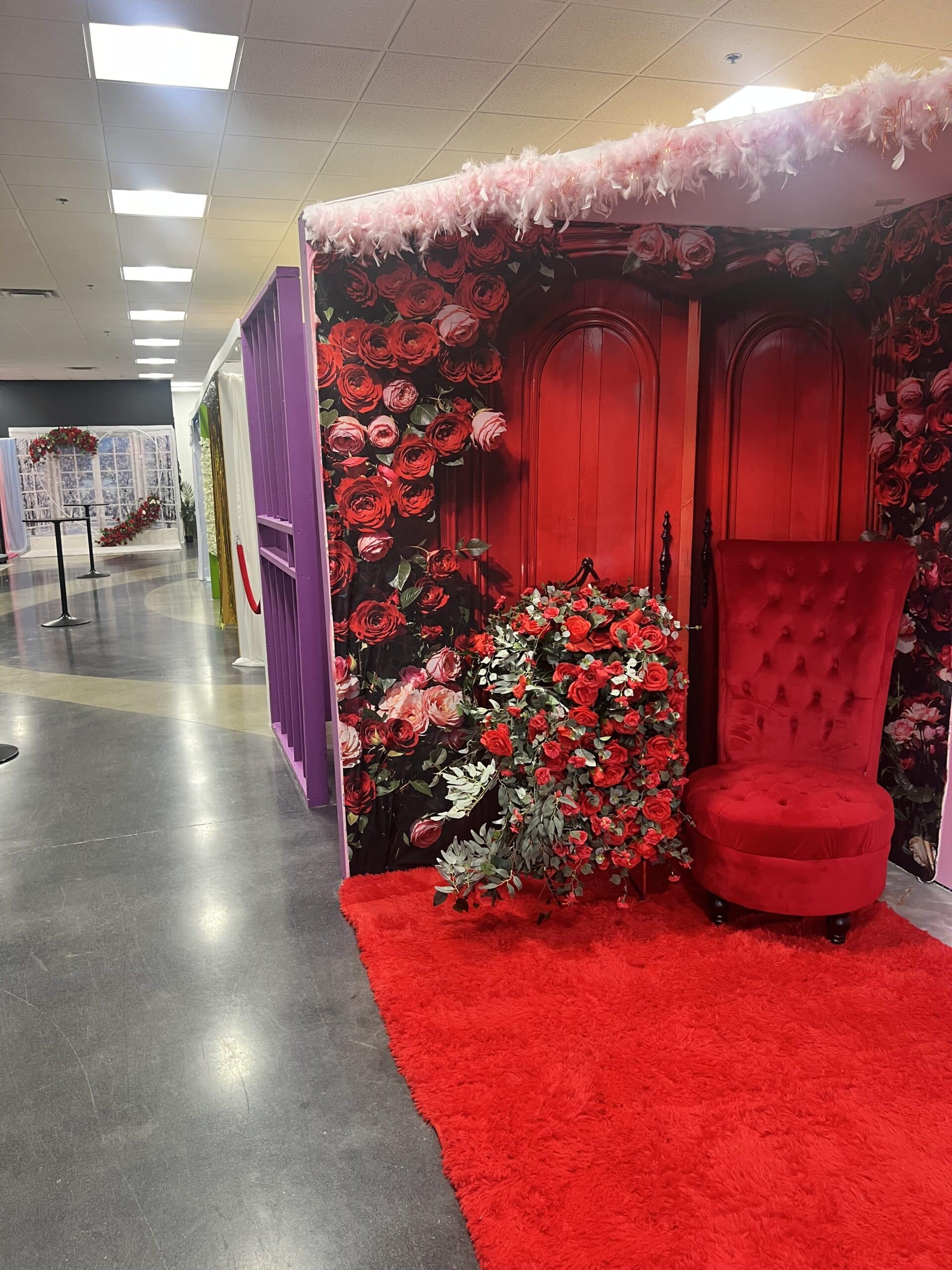 A vibrant holiday-themed indoor display featuring a large red armchair beside a lush bouquet of roses, set against a backdrop of vibrant red doors and festive garlands.