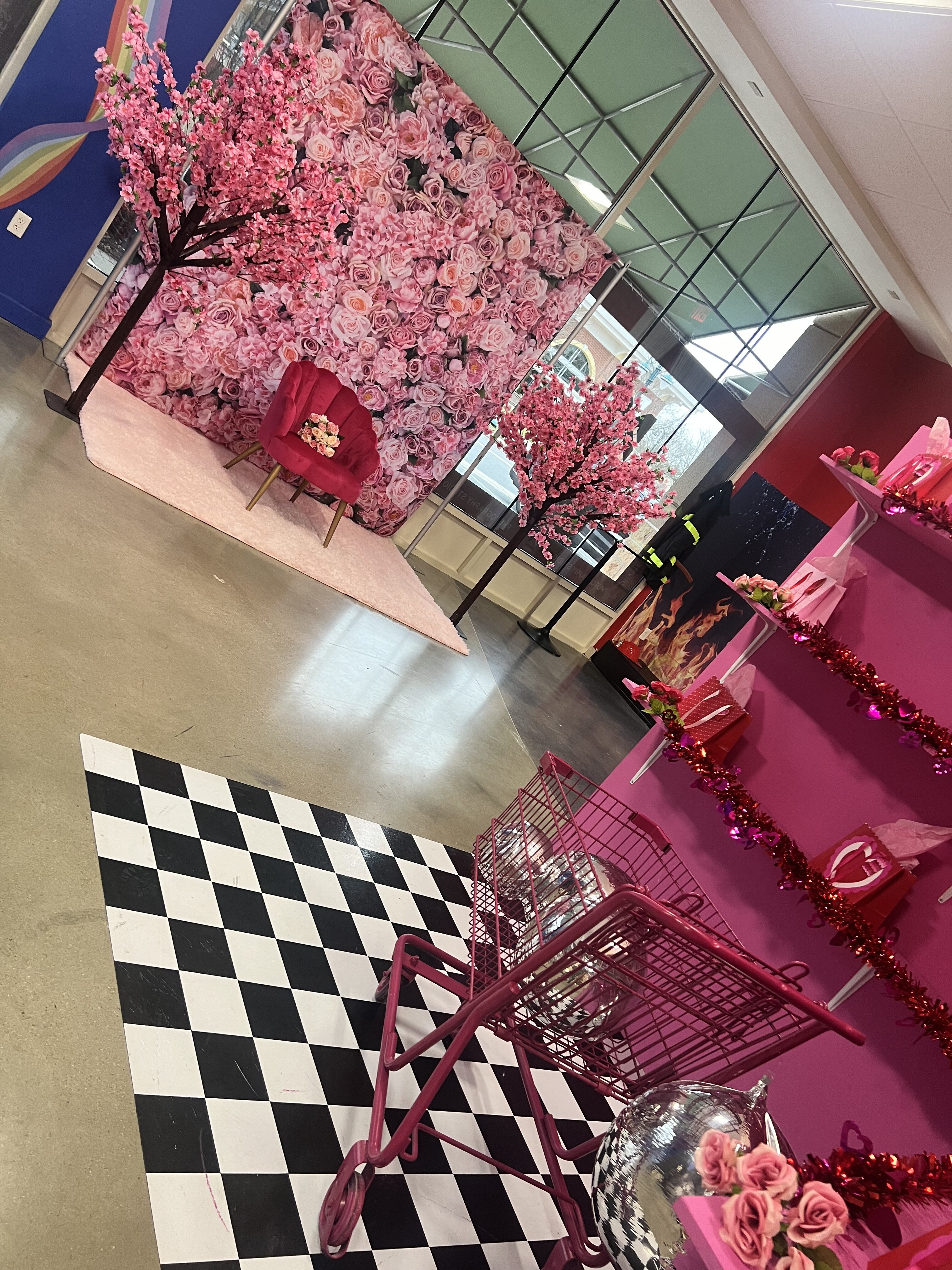 A vibrant pink-themed display area with floral decorations and a checkered black and white floor. a pink armchair and shopping carts add to the lively and stylish setup.