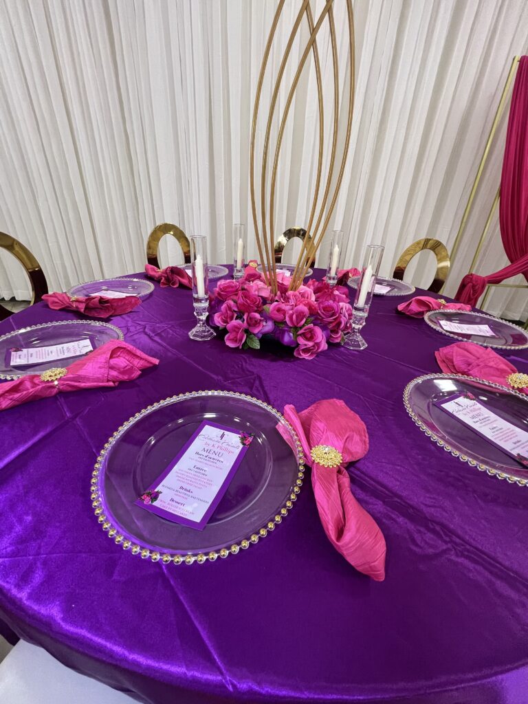 A round table with a bright purple tablecloth is elegantly set for a formal event. Each place setting includes a clear plate adorned with a menu, and a pink napkin styled with a gold napkin ring. The centerpiece features pink and purple flowers and five white candles.