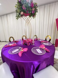 A round table is draped in a purple satin tablecloth with pink napkins folded on clear glass plates. The table centerpiece is an elegant gold stand with lush green leaves and vibrant pink and purple flowers. Six gold-colored chairs with white seats surround the table.