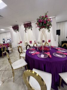 A banquet hall with white walls and marble floors is decorated for an event. A round table features a purple tablecloth, gold chairs, and a tall floral centerpiece. The background has white drapery, a couch area with pink pillows, and hanging floral arrangements.