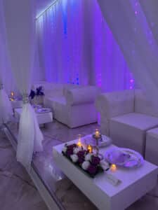 A luxurious room with a plush, white interior, featuring cushioned sofas and white drapes. The room is adorned with purple and white ambient lighting, as well as floral arrangements and lit candles on the tables, creating an elegant and serene atmosphere.