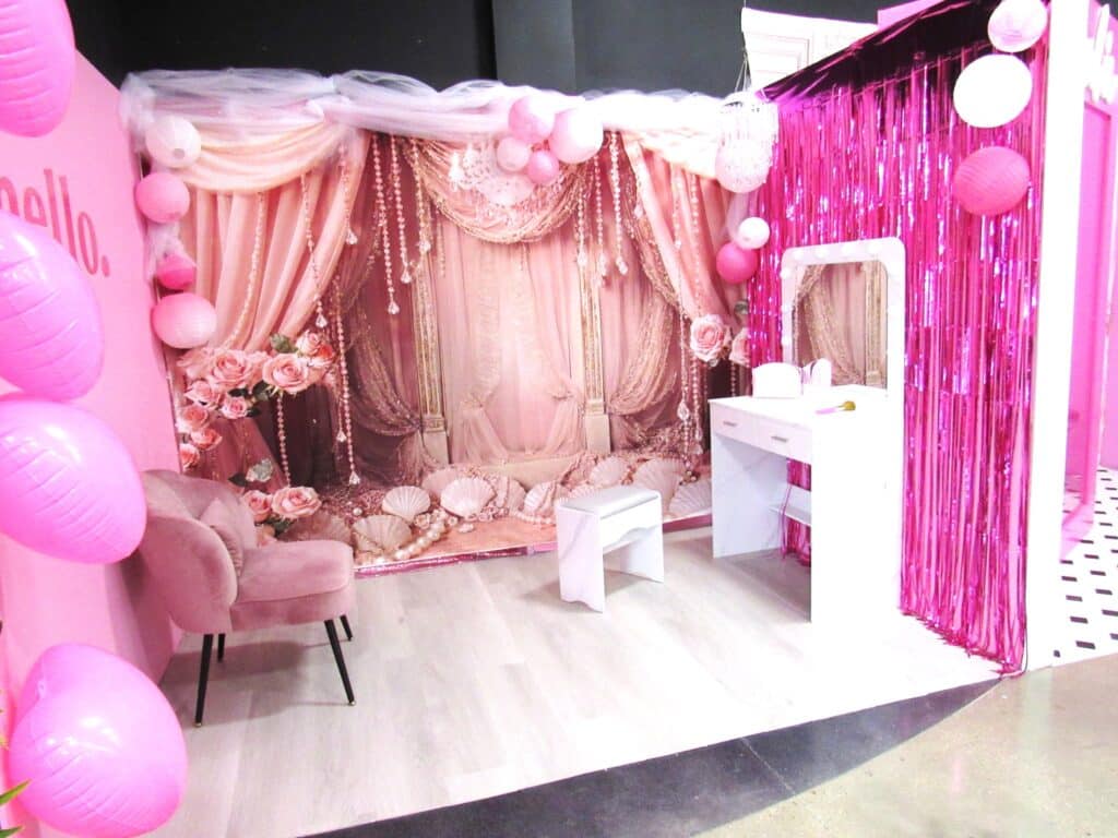 A room decorated with pink and white accents features a vanity table with a mirror, a pink chair, and a draped, ornate backdrop with flowers. Pink balloons and metallic pink streamers add a festive touch. The floor is light wood with black and white tiles on the side.