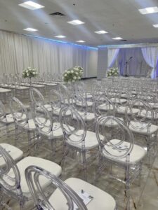 A room arranged for a wedding ceremony with rows of transparent chairs featuring white cushions. The aisle is decorated with large white floral arrangements, and a decorated arch with drapes and fairy lights is set up at the front, with a microphone stand nearby.